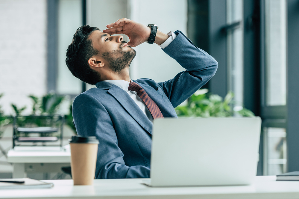 Man in suit sitting at his desk with his eyes closed and hand on his forehead